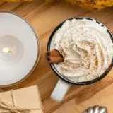 Taza de chocolate caliente con abundante crema de leche, canela y una rama de canela. Una vela encendida y un regalo completan la escena acogedora.