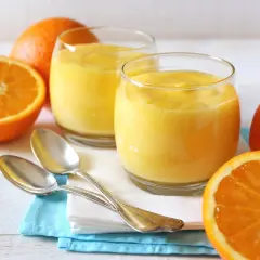 Dos vasos de crema de naranja, naranjas frescas y cucharas plateadas sobre servilletas.