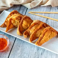 Crujientes wan tun frito dorados en una bandeja blanca, acompañados de salsa agridulce y palillos.