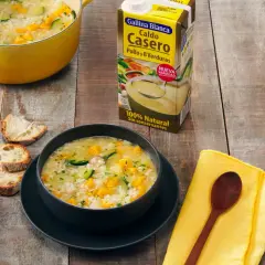 Un tazón humeante de sopa de arroz con calabacín y zanahoria, junto a pan rústico y un cartón de caldo Gallina Blanca.