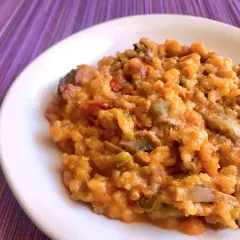 Un cremoso risotto de calabaza con trozos de champiñones y pimientos, servido en un plato blanco sobre un mantel morado.