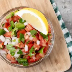 Un tazón de pico de gallo fresco, con tomate, cebolla, pimiento verde y cilantro, adornado con una rodaja de limón.