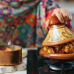 Una mano levanta la tapa de un tajín, revelando un guiso humeante. Descubre qué es el tajín, un plato marroquí.
