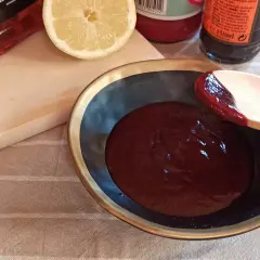 Cuchara de madera prueba la deliciosa salsa barbacoa casera en un cuenco. Limón y botellas de ingredientes al fondo.