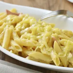 Un plato blanco ovalado lleno de macarrones con queso cremosos y trozos de tocino. Un tenedor plateado descansa sobre el borde del plato.