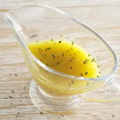 Vinagreta de mostaza dorada con hierbas verdes picadas, servida en una salsera de cristal sobre una mesa de madera.