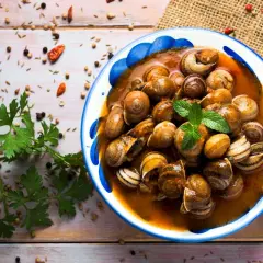 Un plato de caracoles en salsa, adornado con menta fresca, rodeado de especias y perejil sobre una mesa de madera.