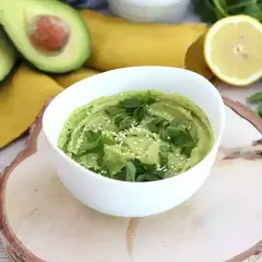 Un cremoso hummus de aguacate, decorado con perejil fresco y semillas de sésamo, junto a aguacates cortados y un limón.