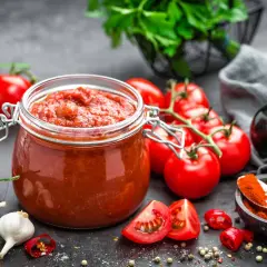 Frasco de vidrio lleno de vibrante salsa de tomate casera, rodeado de tomates frescos, ajos, chiles y especias.