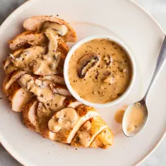 Pechuga de pollo en rodajas cubierta con una cremosa salsa de setas, servida con un tazón de la misma salsa y una cuchara.