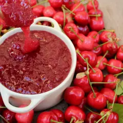 Cuchara vierte mermelada de cerezas casera en un cuenco blanco, rodeado de cerezas frescas y hojas verdes.