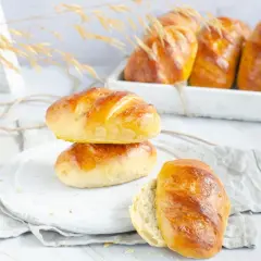 Dos panes de leche dorados apilados, uno abierto, con más panes en una bandeja al fondo.
