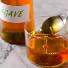 Cuchara con sirope de agave cayendo en un vaso, junto a una botella etiquetada.
