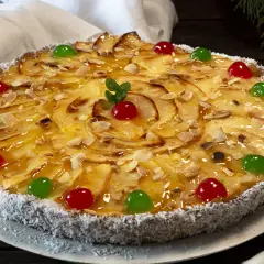 Tarta de manzana con crema pastelera, decorada con almendras laminadas, cerezas rojas y verdes, y coco rallado en el borde.