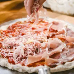 Mano esparciendo queso rallado sobre una masa de pizza casera con jamón y salsa de tomate.