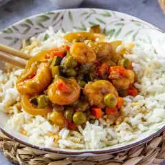 Un plato de arroz blanco cubierto con gambas en salsa agridulce y verduras. Los palillos descansan sobre el borde del cuenco.