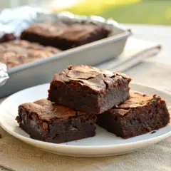 Deliciosos trozos de brownie sin gluten apilados en un plato blanco. En el fondo, más brownies en un molde.