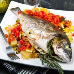 Dorada al horno con verduras asadas, servida en plato blanco con tenedor y romero fresco.