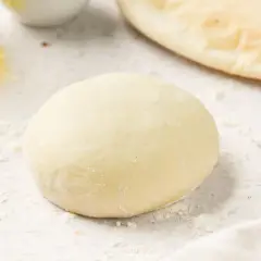 Masa de pizza sin levadura lista para estirar. Harina esparcida, aceite y un trozo de pan plano al fondo.