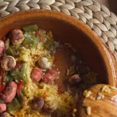 Un plato de barro con arroz, salchichas, pimientos verdes y rojos. Un cucharón de madera reposa en el borde.