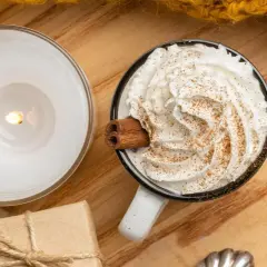 Taza de chocolate caliente con abundante crema de leche, canela y una rama de canela. Una vela encendida y un regalo completan la escena acogedora.