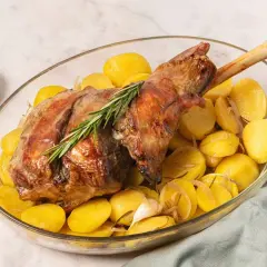 Pierna de cordero al horno dorada y jugosa, acompañada de patatas asadas y romero en una fuente de cristal.