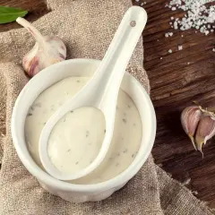 Crema de salsa de ajo en un cuenco blanco con una cuchara, rodeada de ajos y hojas verdes sobre una mesa de madera rústica.