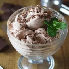 Delicioso helado de chocolate casero en copa, adornado con virutas de chocolate y una hoja de menta. A su lado, trozos de chocolate negro y una cuchara.