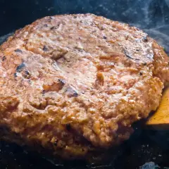Jugosa hamburguesa de ternera dorándose en sartén, con vapor ascendiendo y una espátula de madera lista para darle la vuelta.