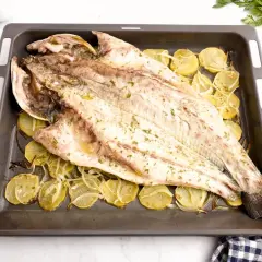 Corvina al horno, entera y dorada, reposa sobre rodajas de patata y cebolla en una bandeja.