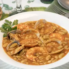 Plato de verdinas con almejas y gambas, servido en un cuenco blanco con perejil. Acompañado de pan en una cesta.