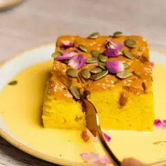 Un trozo de bizcocho de calabaza, decorado con semillas de calabaza y pétalos de rosa, servido en una crema amarilla.