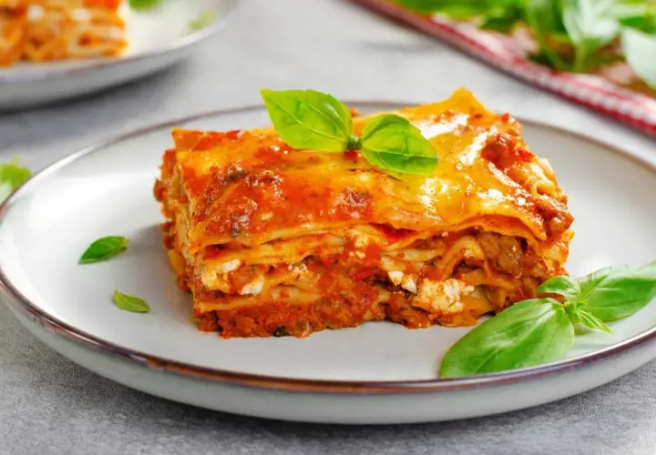 Una porción de lasaña de carne, con capas de pasta, salsa y queso, decorada con hojas de albahaca fresca.