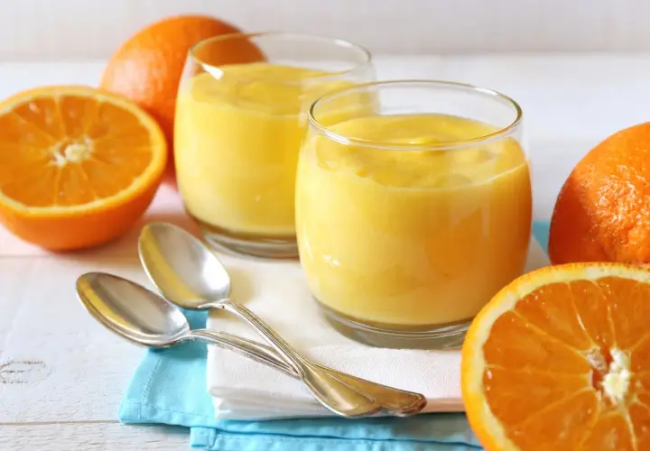 Dos vasos de crema de naranja, naranjas frescas y cucharas plateadas sobre servilletas.