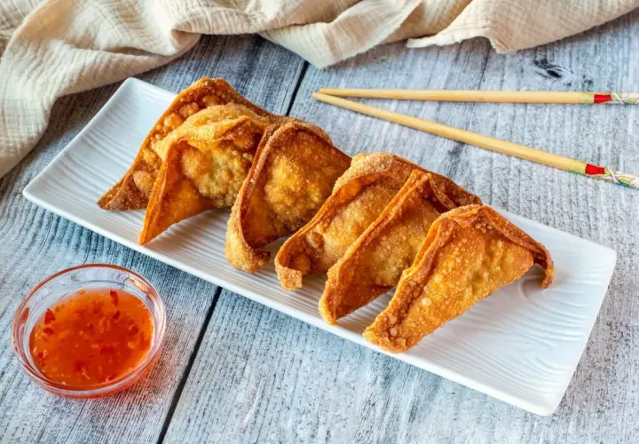 Crujientes wan tun frito dorados en una bandeja blanca, acompañados de salsa agridulce y palillos.