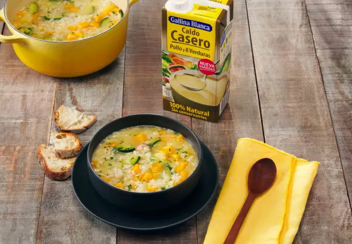 Un tazón humeante de sopa de arroz con calabacín y zanahoria, junto a pan rústico y un cartón de caldo Gallina Blanca.