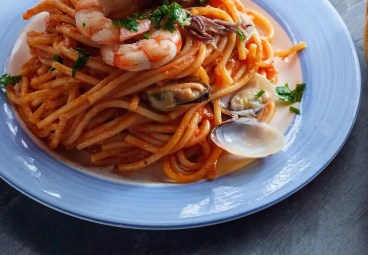 Espaguetis a la marinera con gambas, mejillones y almejas en una salsa roja, servidos en un plato azul.