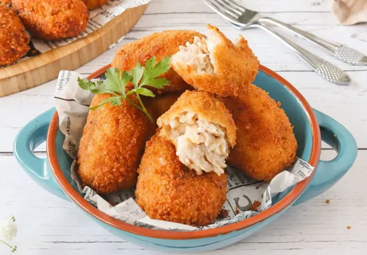 Croquetas de atún doradas y crujientes, servidas en un cuenco azul. Una está partida, mostrando su cremoso interior.