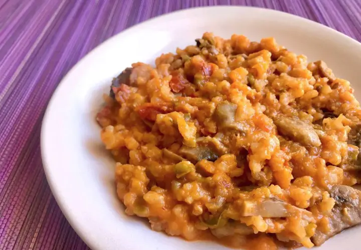 Un cremoso risotto de calabaza con trozos de champiñones y pimientos, servido en un plato blanco sobre un mantel morado.