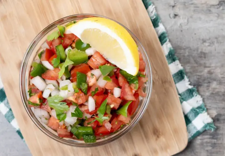 Un tazón de pico de gallo fresco, con tomate, cebolla, pimiento verde y cilantro, adornado con una rodaja de limón.