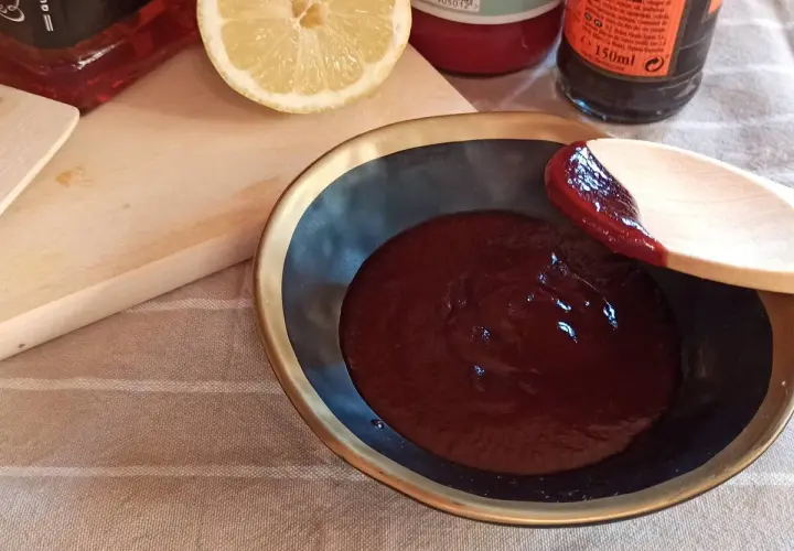 Cuchara de madera prueba la deliciosa salsa barbacoa casera en un cuenco. Limón y botellas de ingredientes al fondo.