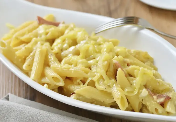 Un plato blanco ovalado lleno de macarrones con queso cremosos y trozos de tocino. Un tenedor plateado descansa sobre el borde del plato.