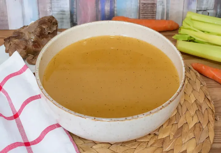 Un tazón de caldo de carne humeante, con un trozo de carne y verduras frescas al lado.