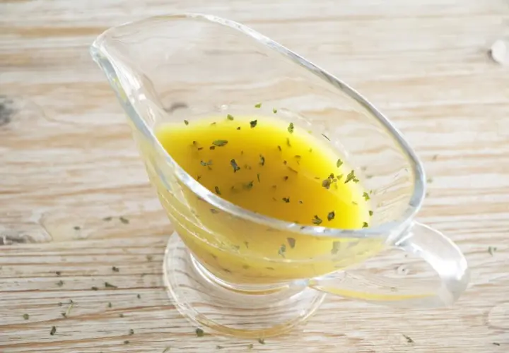 Vinagreta de mostaza dorada con hierbas verdes picadas, servida en una salsera de cristal sobre una mesa de madera.