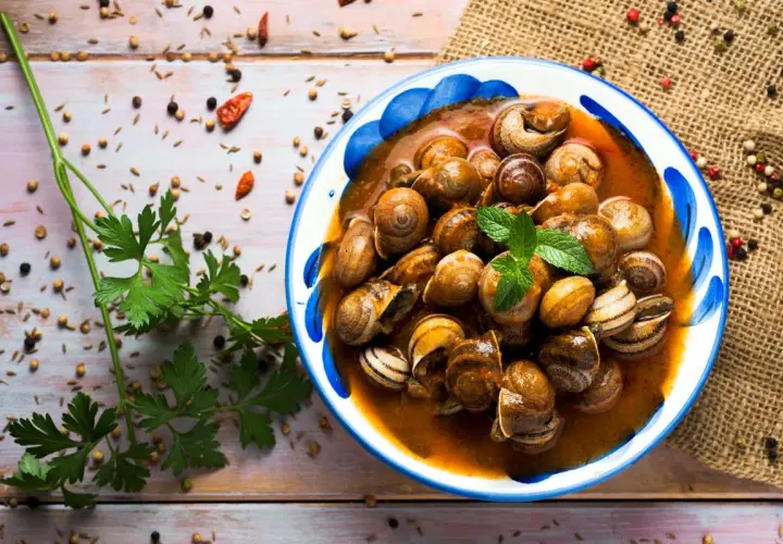 Un plato de caracoles en salsa, adornado con menta fresca, rodeado de especias y perejil sobre una mesa de madera.