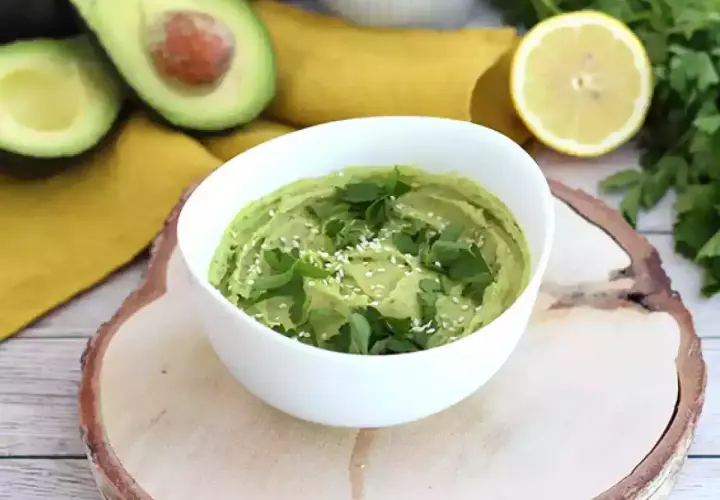 Un cremoso hummus de aguacate, decorado con perejil fresco y semillas de sésamo, junto a aguacates cortados y un limón.