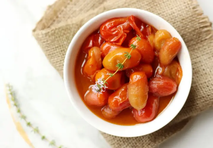 Un tazón blanco lleno de tomates cherry confitados, con una ramita de tomillo y una flor morada.