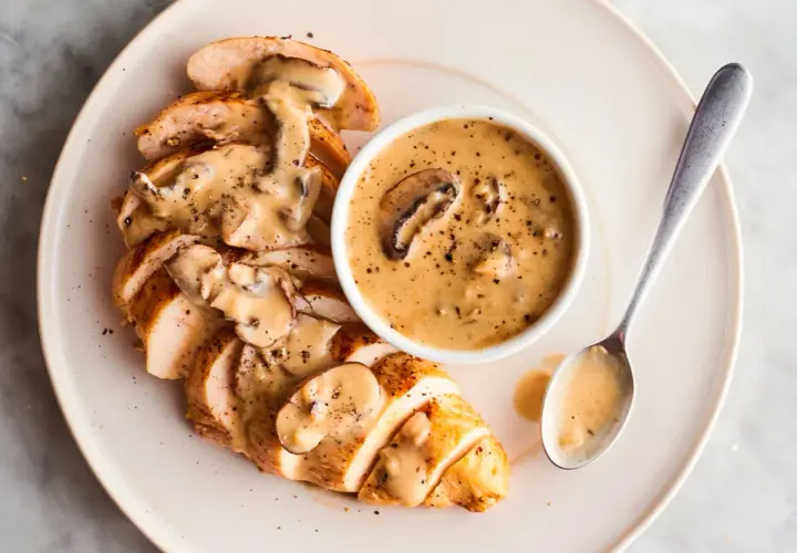 Pechuga de pollo en rodajas cubierta con una cremosa salsa de setas, servida con un tazón de la misma salsa y una cuchara.