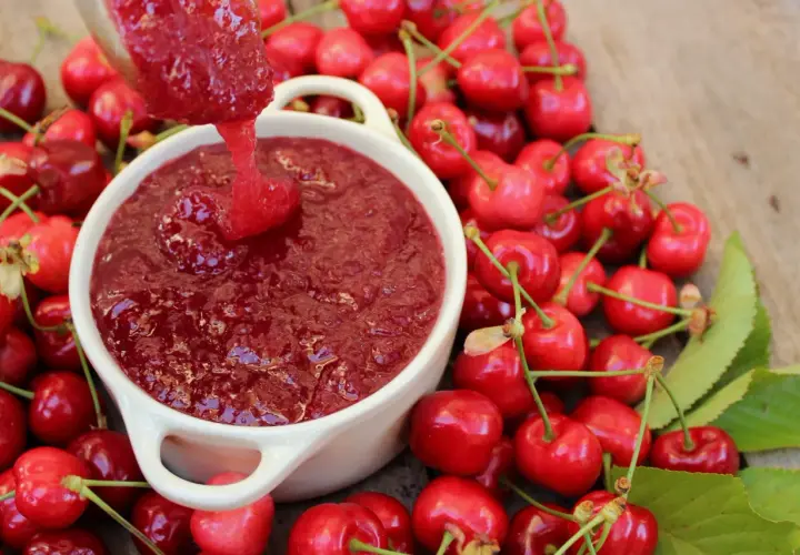 Cuchara vierte mermelada de cerezas casera en un cuenco blanco, rodeado de cerezas frescas y hojas verdes.