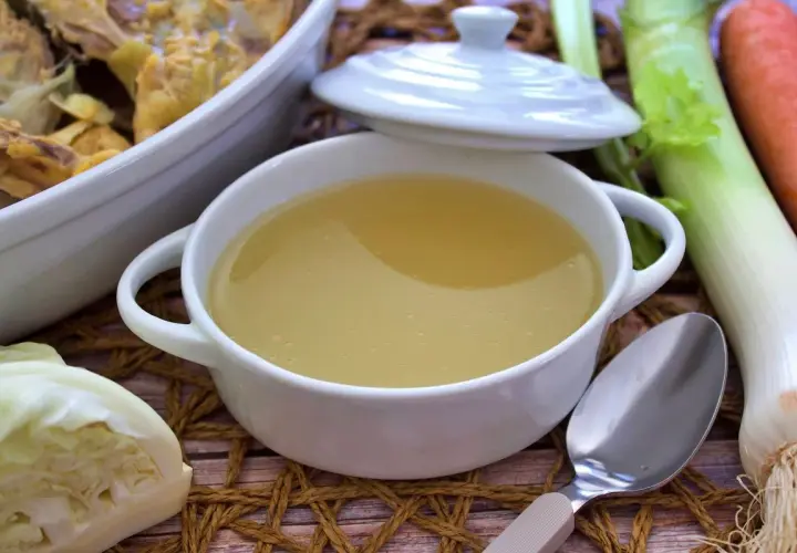 Un tazón humeante de caldo de pollo casero, listo para reconfortar. Verduras frescas como puerro, zanahoria y repollo acompañan.